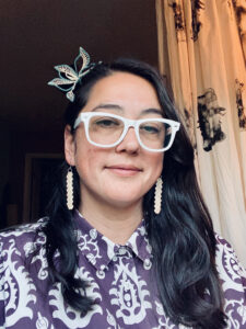 Headshot of Lehua M. Taitano wearing white-framed glasses and a blue and white patterned shirt.