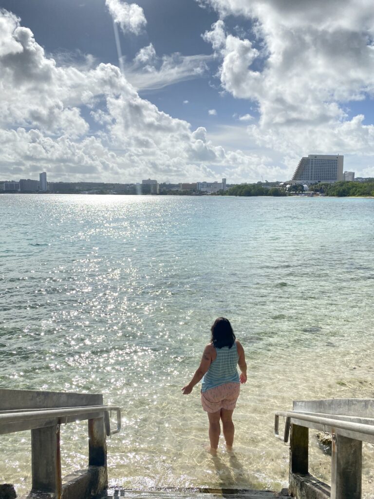 A woman on the beach looking out across the teal waters under a blue sky with clouds.